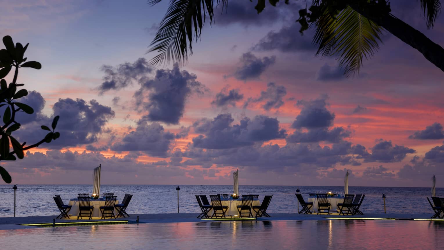 Tables with chairs between infinity pool and ocean at dusk with palm tree