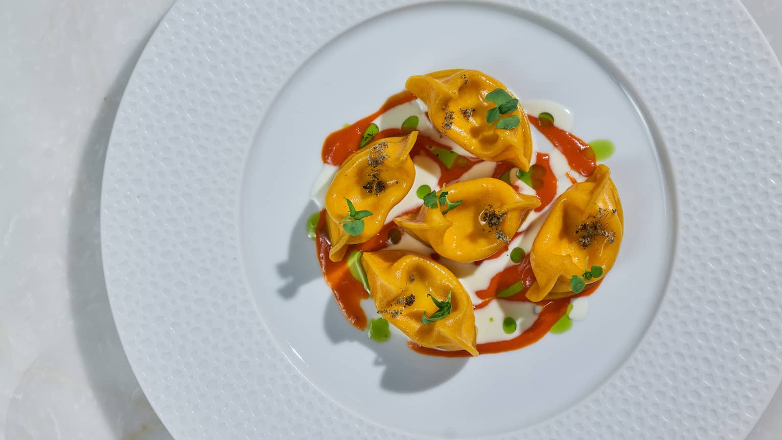 A white plate with a textured border holds five orange food items topped with green garnish and drizzled with a red sauce.