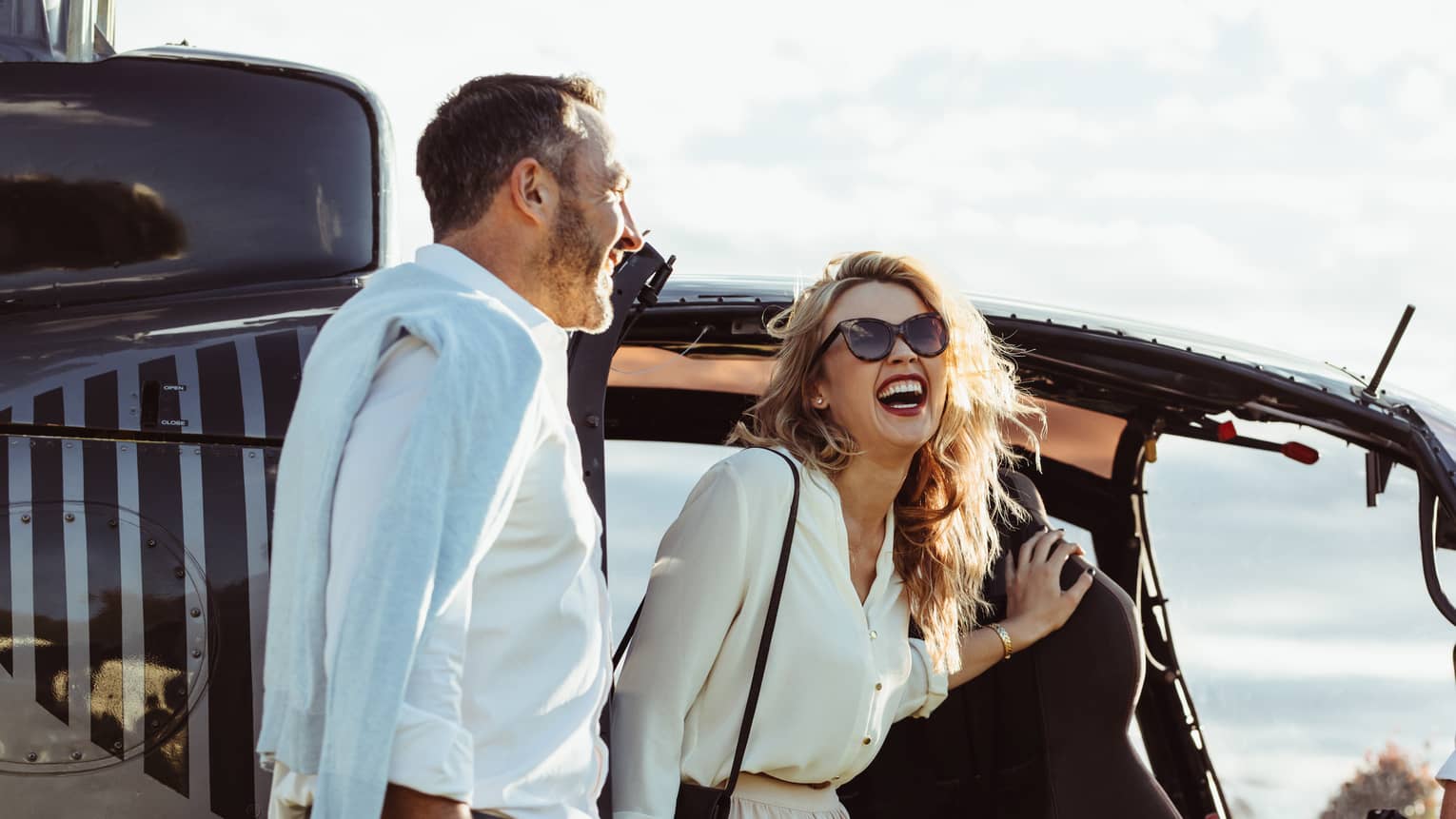 A man and woman laughing while getting out of a helicopter with the pilot smiling nearby.