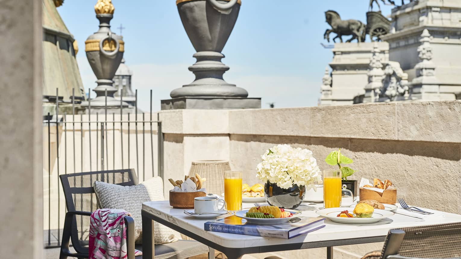 Breakfast setting on a luxury hotel terrace in Madrid with a table featuring orange juice, pastries, flowers and a book, overlooking historic architecture 