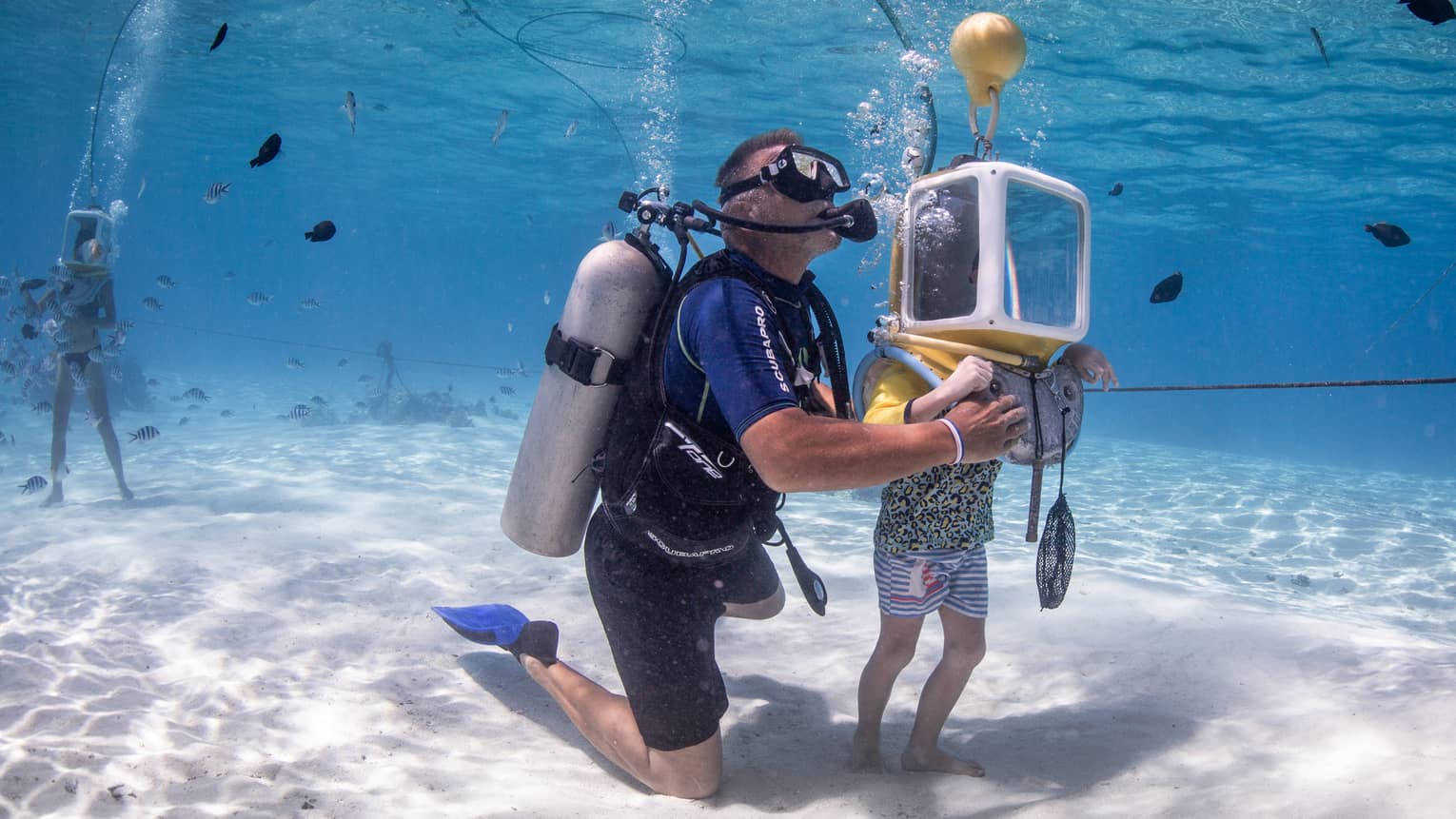 An instructor in scuba gear helps a child in a glass diving helmet traverse the ocean floor as fish swim all around them.