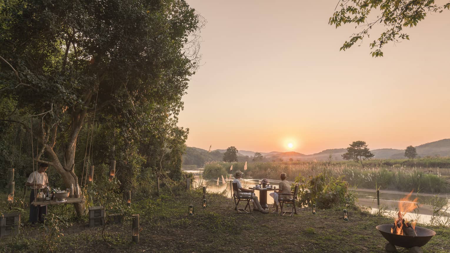 Outdoor riverside dining setup at sunset with a campfire and mountain views