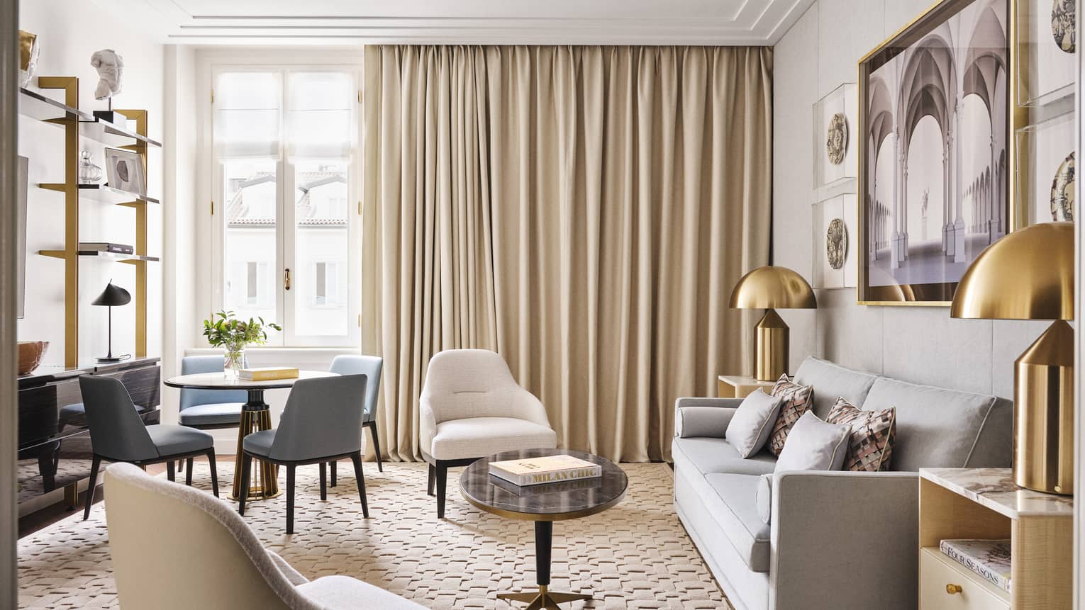 Contemporary living room of a hotel suite, featuring grey sofa, brass lamps, two off-white arm chairs and round dining table with four blue chairs