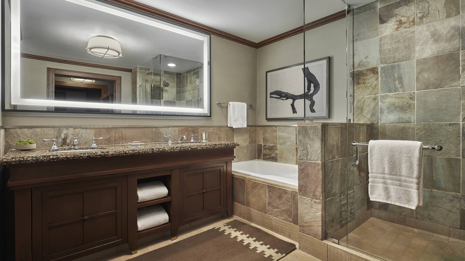 A tiled bathroom, with a double vanity, large lighted mirror and a shower and tub separated by a glass partition