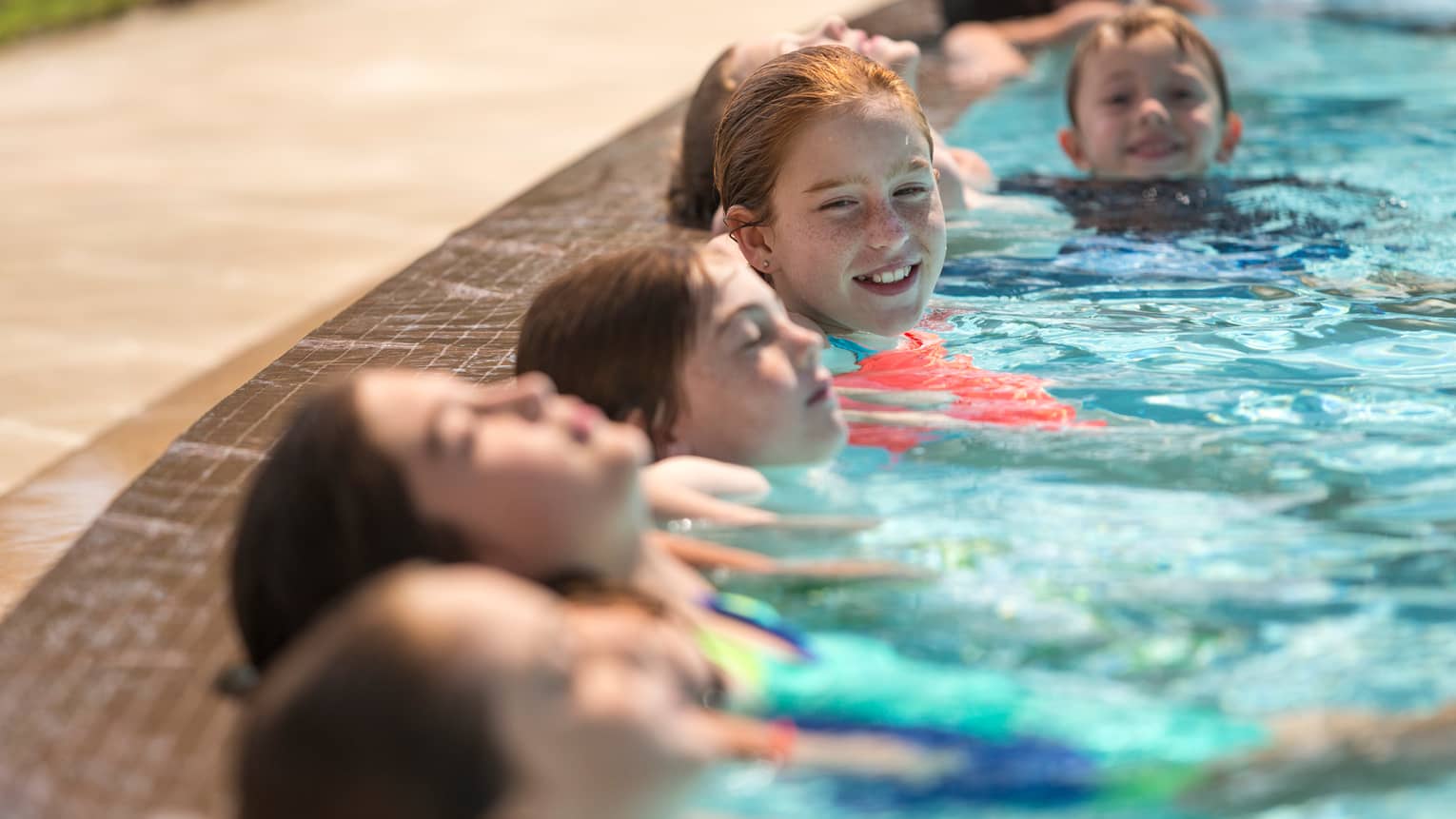 Six children smile, lean their heads against side of swimming pool