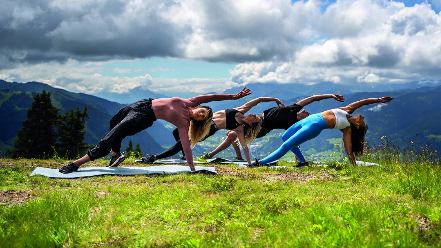 Four people in bending side plank yoga pose on meadow with mountains in backdrop