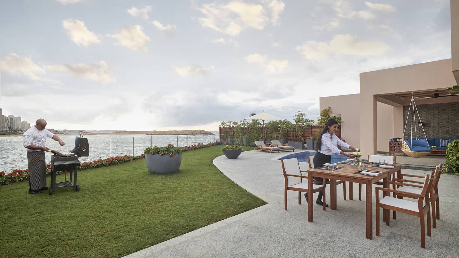 A chef seasons meat on a barbecue near the water’s edge while a server arranges flowers at a table on a patio near a pool.