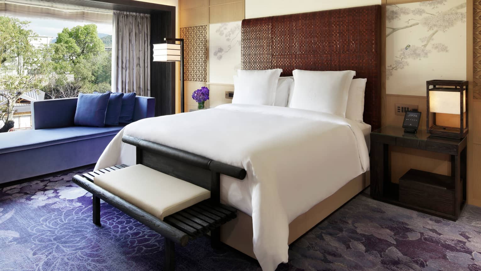 Elegant bedroom with a king bed, a purple chaise lounge, rug and flowers and floor-to-ceiling windows overlooking lush greenery