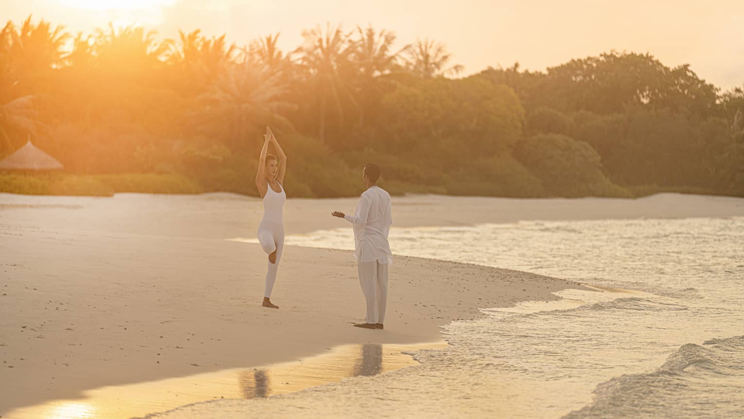 Person practicing yoga on a tropical beach at sunrise with an instructor, surrounded by calm ocean waves and lush greenery in the background