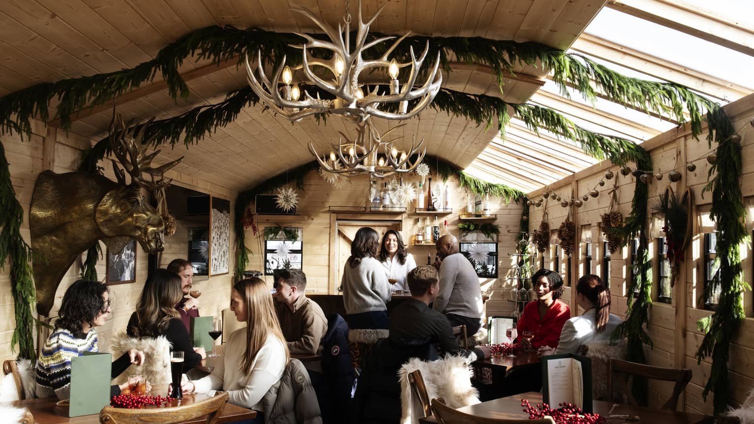 Nordic-style bar with antler chandeliers, wooden ceiling trimmed with greenery and people sitting at various tables