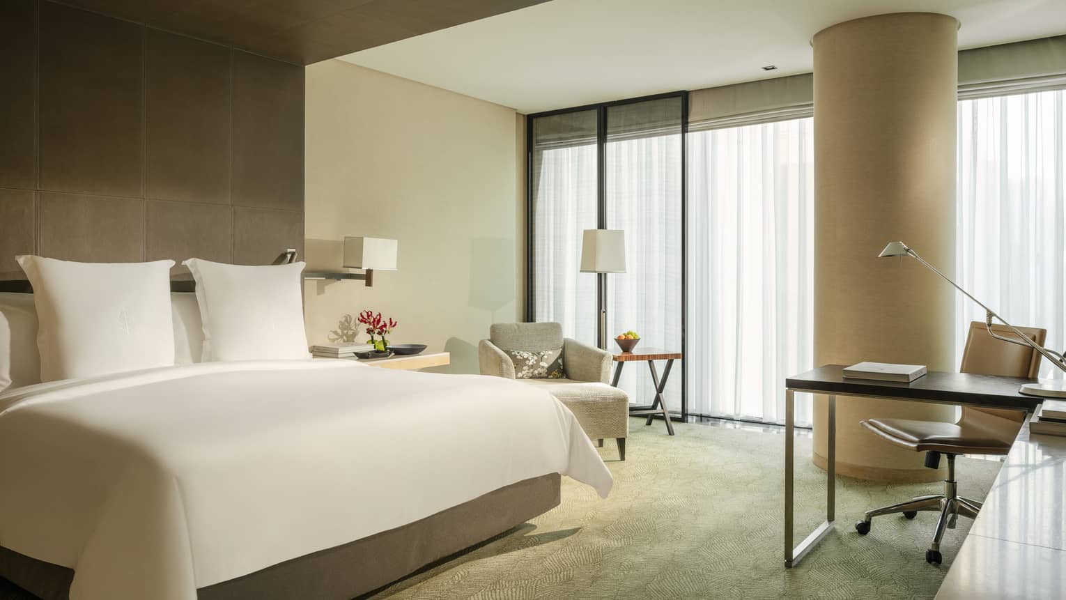 Guest room featuring a king bed, armchair, workspace, side table with red flowers and floor-to-ceiling windows with curtains