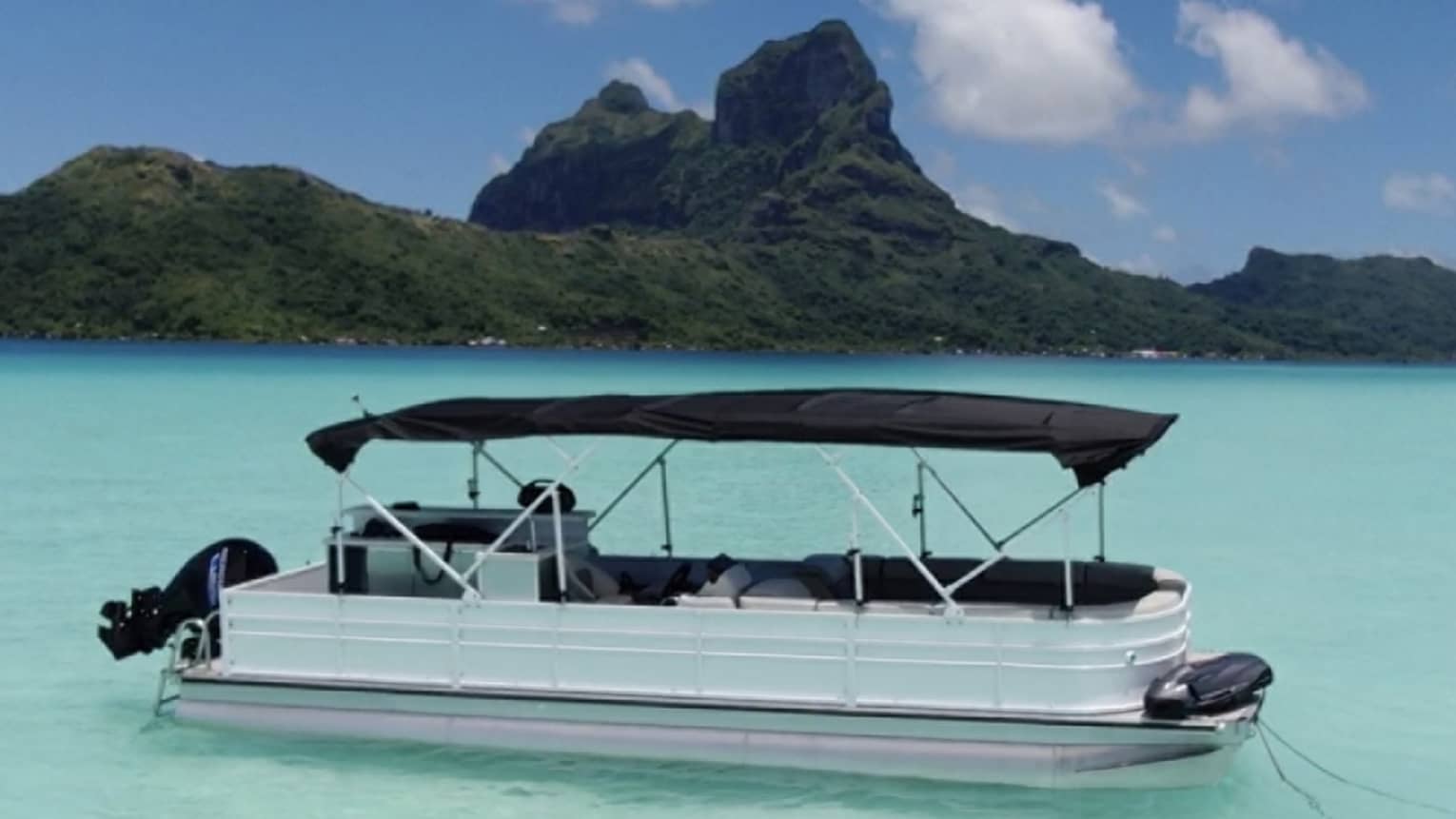 A pontoon boat with a canvas roof anchored in pale turquoise waters; a forested mountain looms behind.
