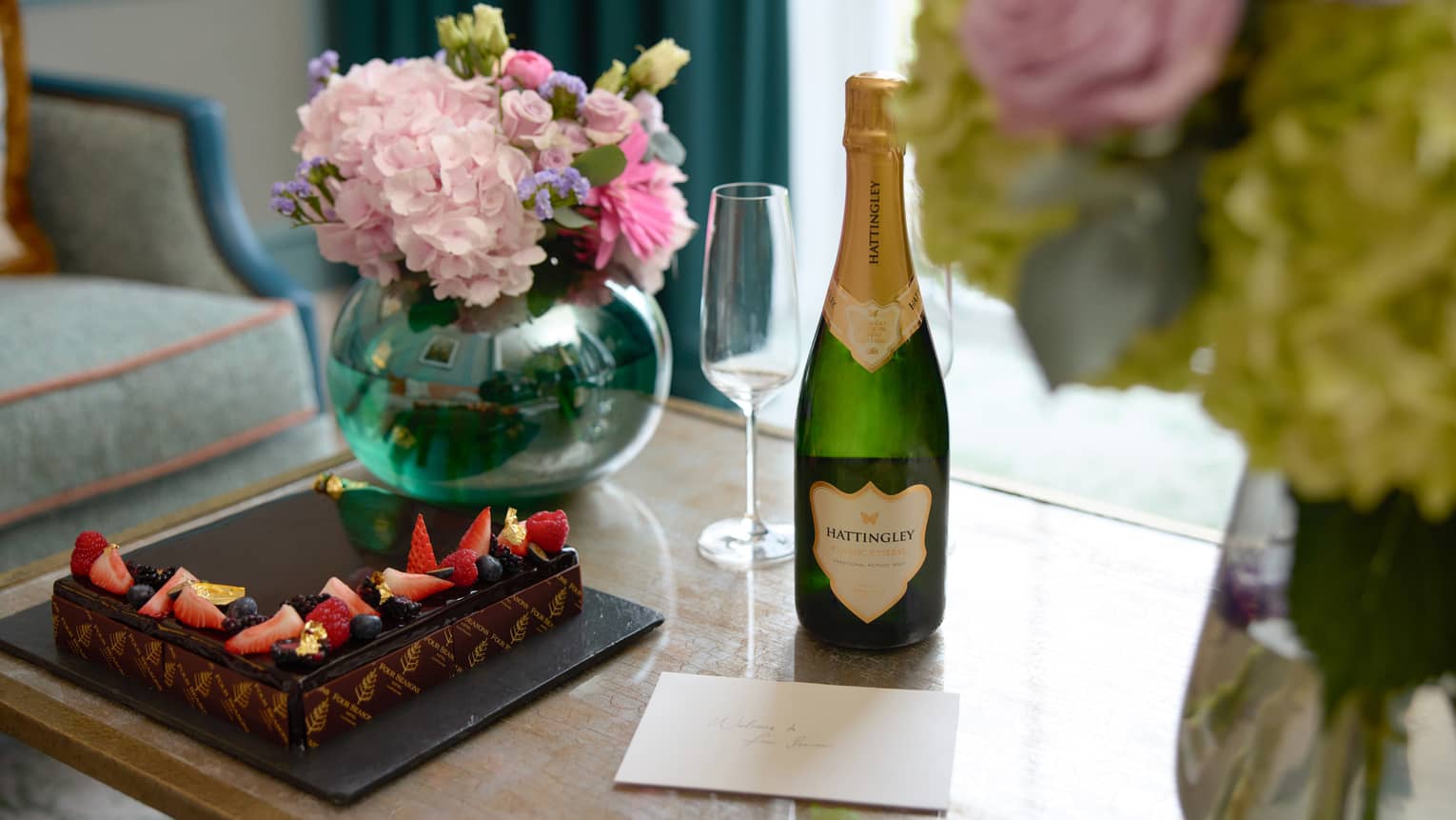 A small cake, bottle of champagne, stemware and floral arrangements sit on a table in a room.