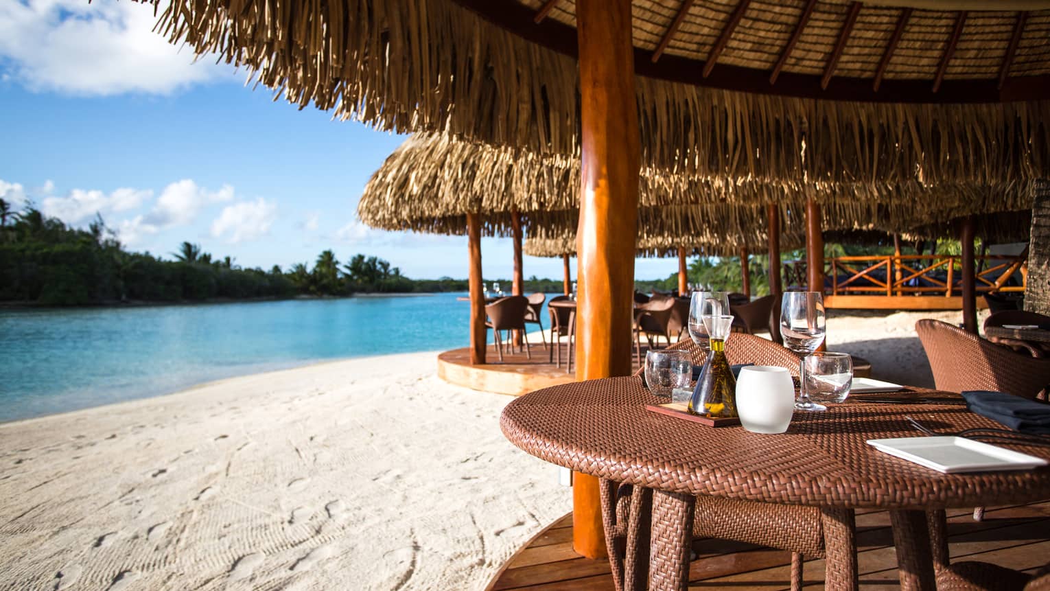 Edge of round rattan patio dining tables under thatched roof pavilion on white sand beach