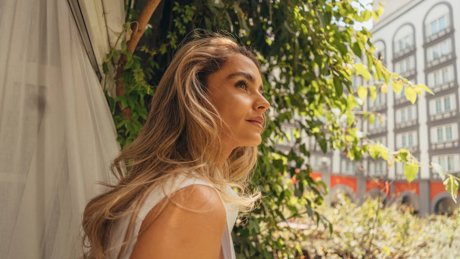A person in an outdoor setting with foliage in the near background and the wall of a building with windows in the far background gazes off camera
