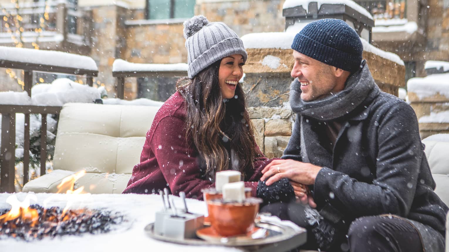 Smiling couple sitting by a firepit in a snowy outdoor setting, bundled in winter coats and hats. Snowflakes fall around them as they enjoy a cosy moment. 