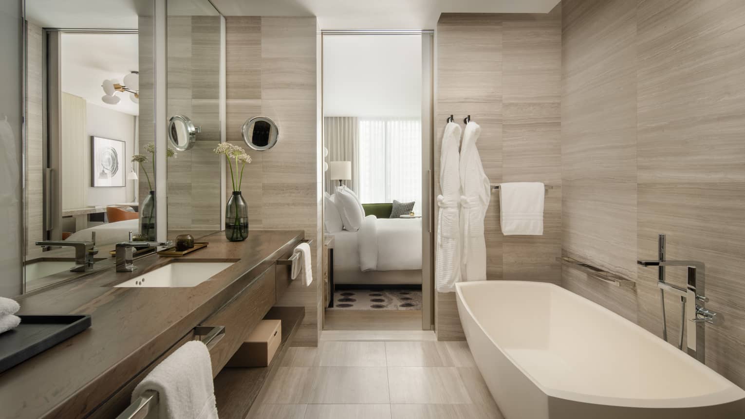 Luxury hotel bathroom with a freestanding tub, spacious vanity, large mirror, and two white bathrobes hanging beside the door