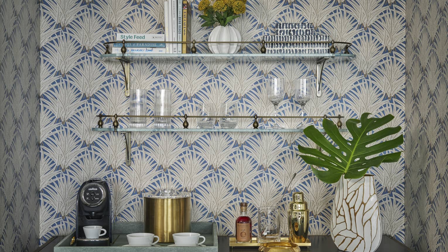 Stylish beverage station with a coffee maker, glass shelves holding cups, glasses, books and a vase and decorative wallpaper with a leaf pattern