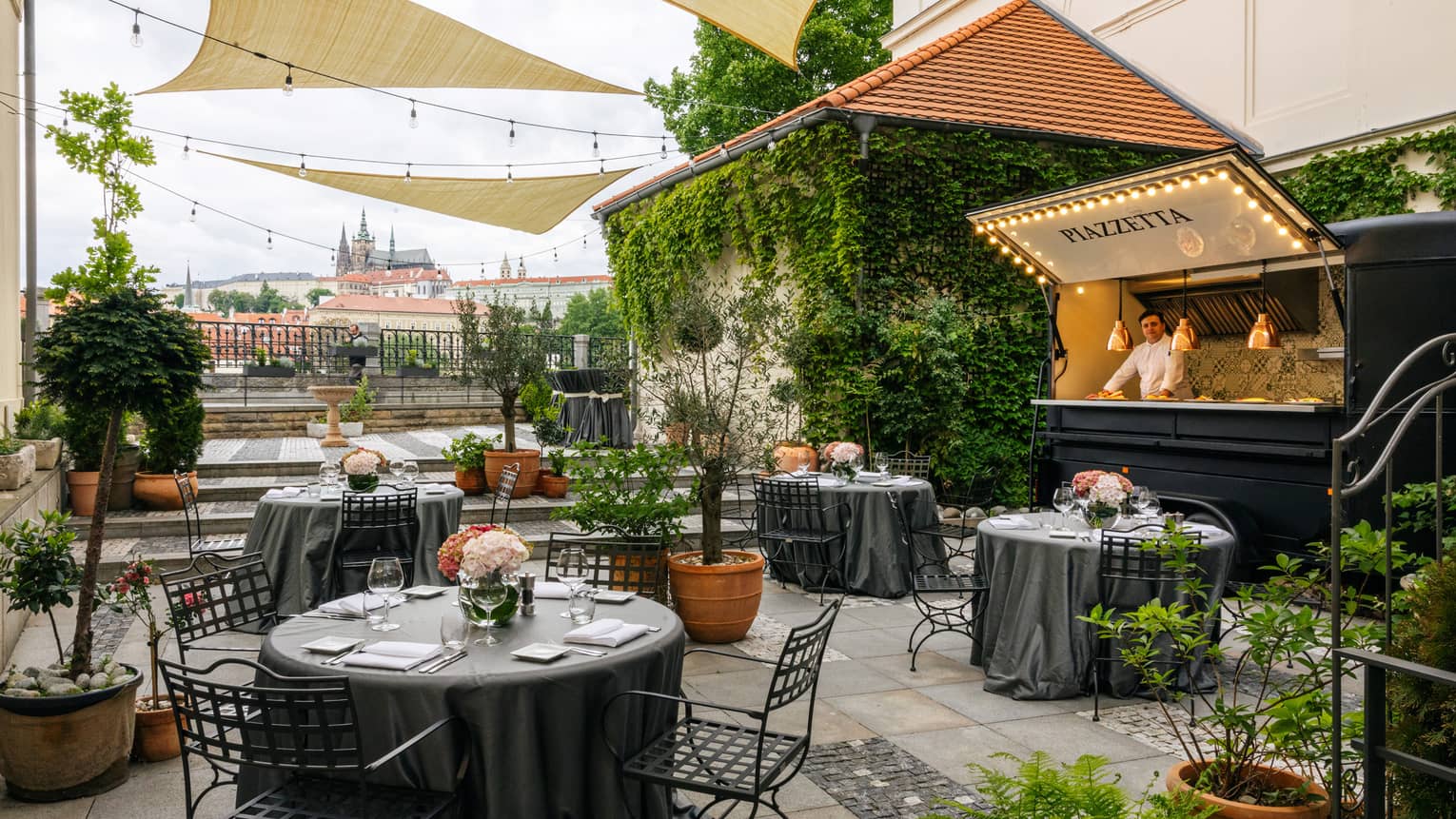 Piazzetta rooftop restaurant with black round iron tables, topiaries, mixologist behind bar, Prague Castle view