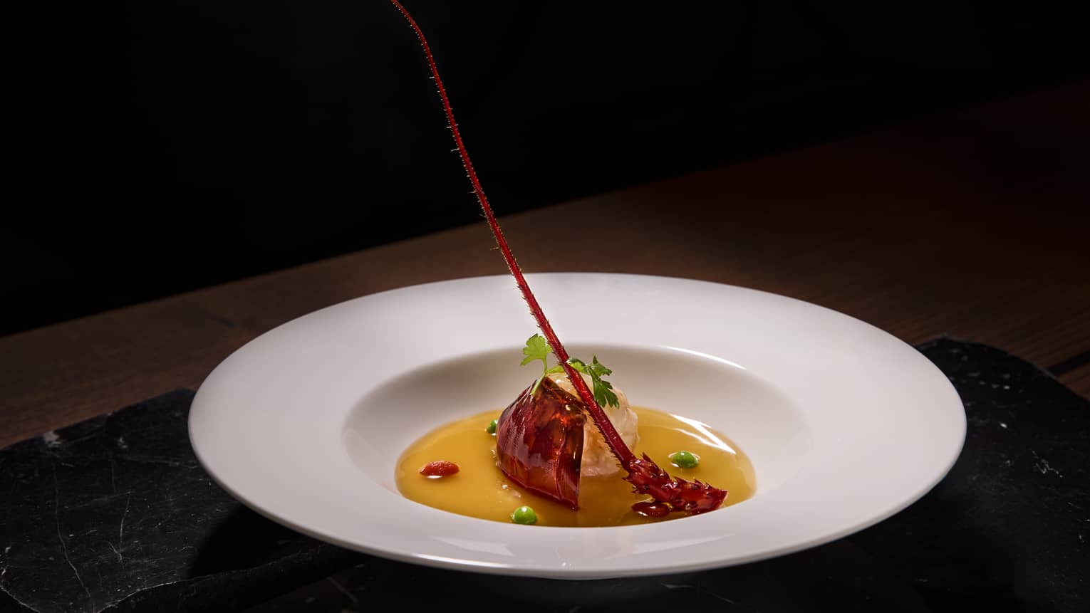 A red food item sits in a dollop of light orange sauce in a shallow white bowl; the food is topped with small pieces of leafy green garnish.