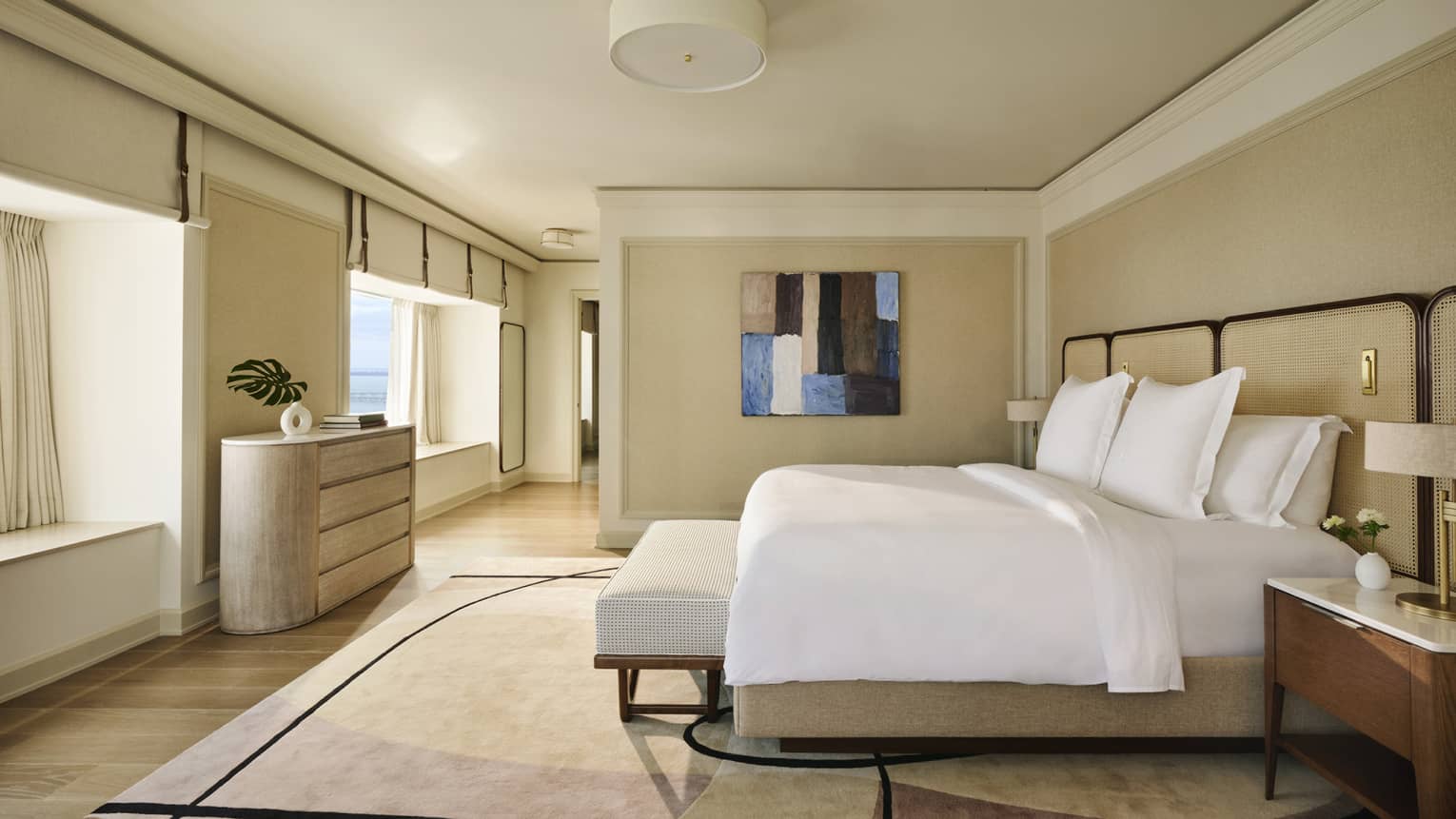 Hotel suite bedroom with king bed, beige walls, abstract artwork and area rug, and windows