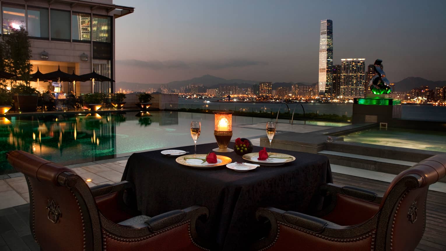 Candle-lit patio dining table by swimming pool at night, skyscrapers and city lights