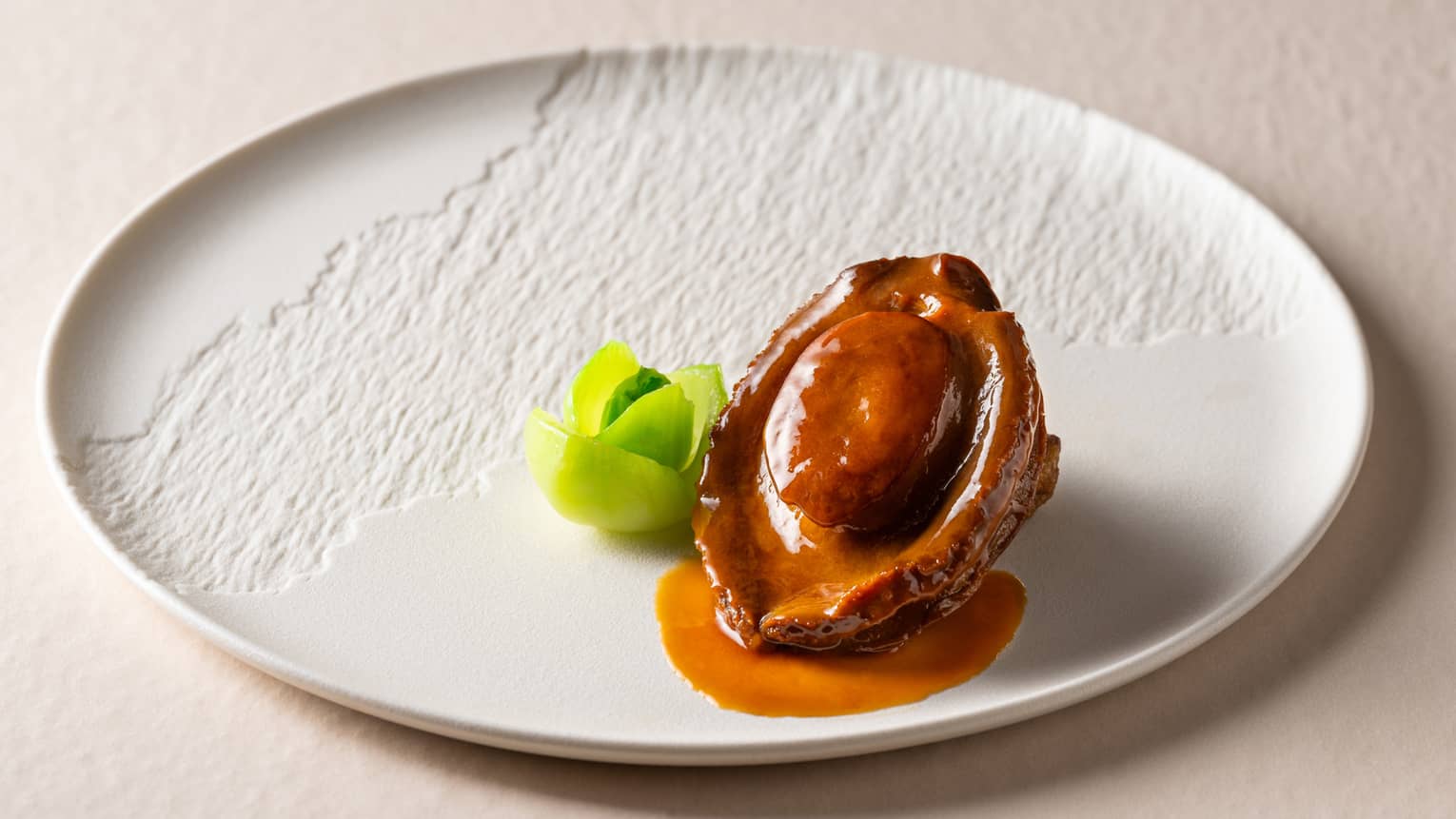 Textured white plate with abalone in sauce and green vegetable garnish