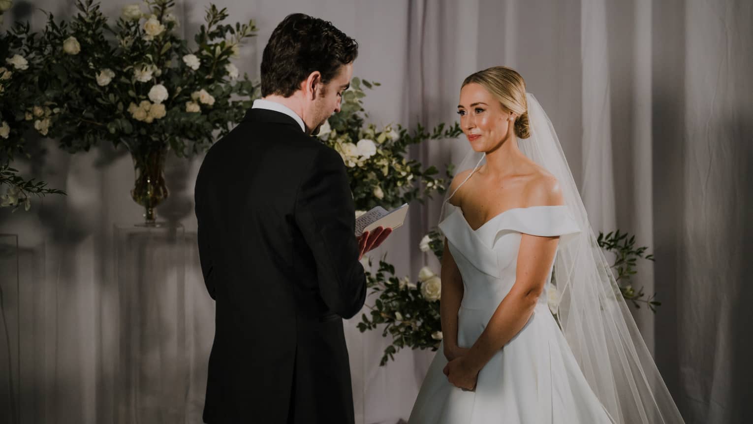 A groom reads from a vow book to a bride at an indoor wedding ceremony