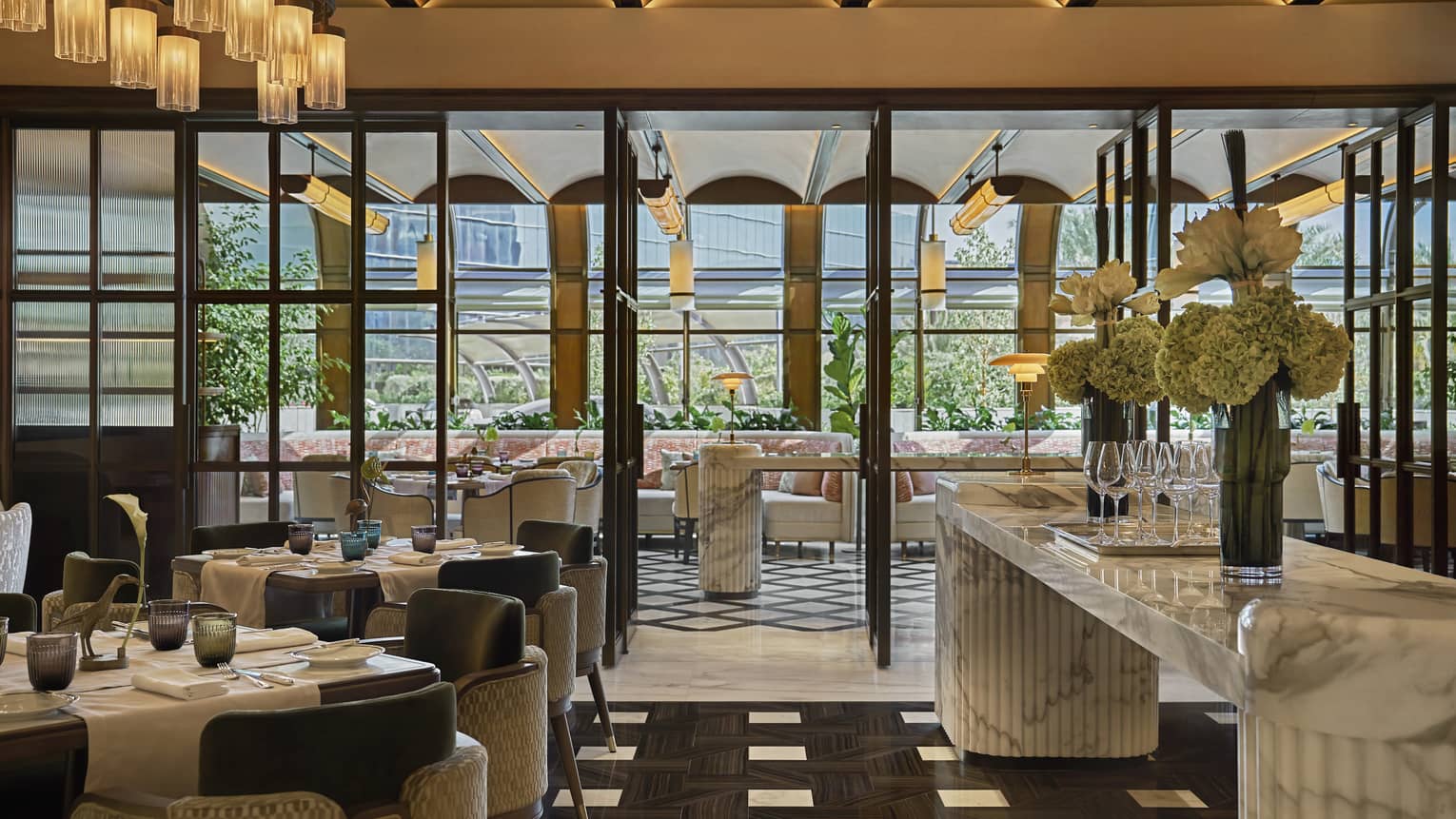 Sun-lit dining restaurant dining room with black-and-white tile flooring, a large white marble counter and floor-to-ceiling glass doors that lead out to a terrace with more seating