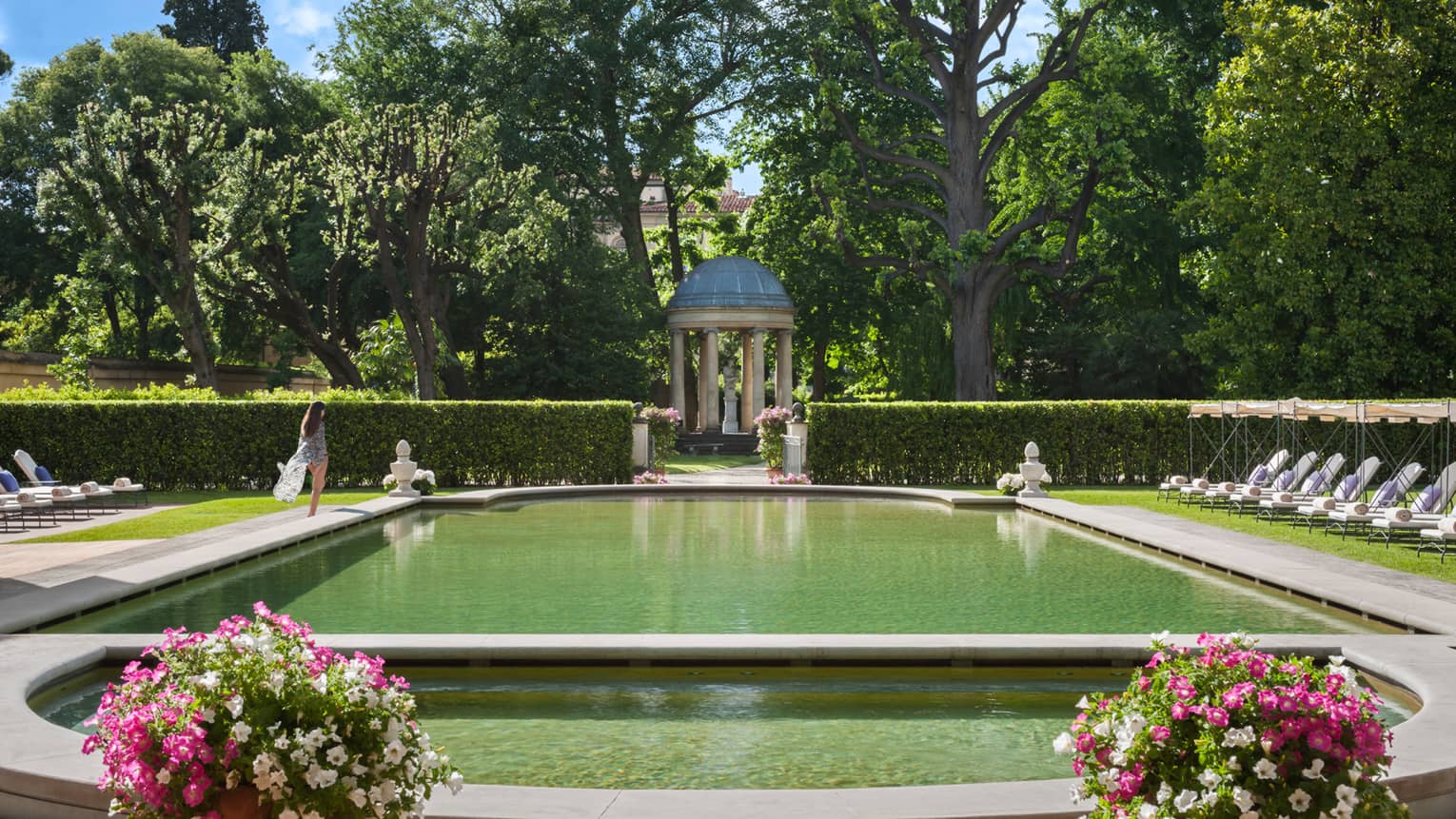 Woman in flowing robe walks past stone ledge of outdoor swimming pool surrounded by gardens