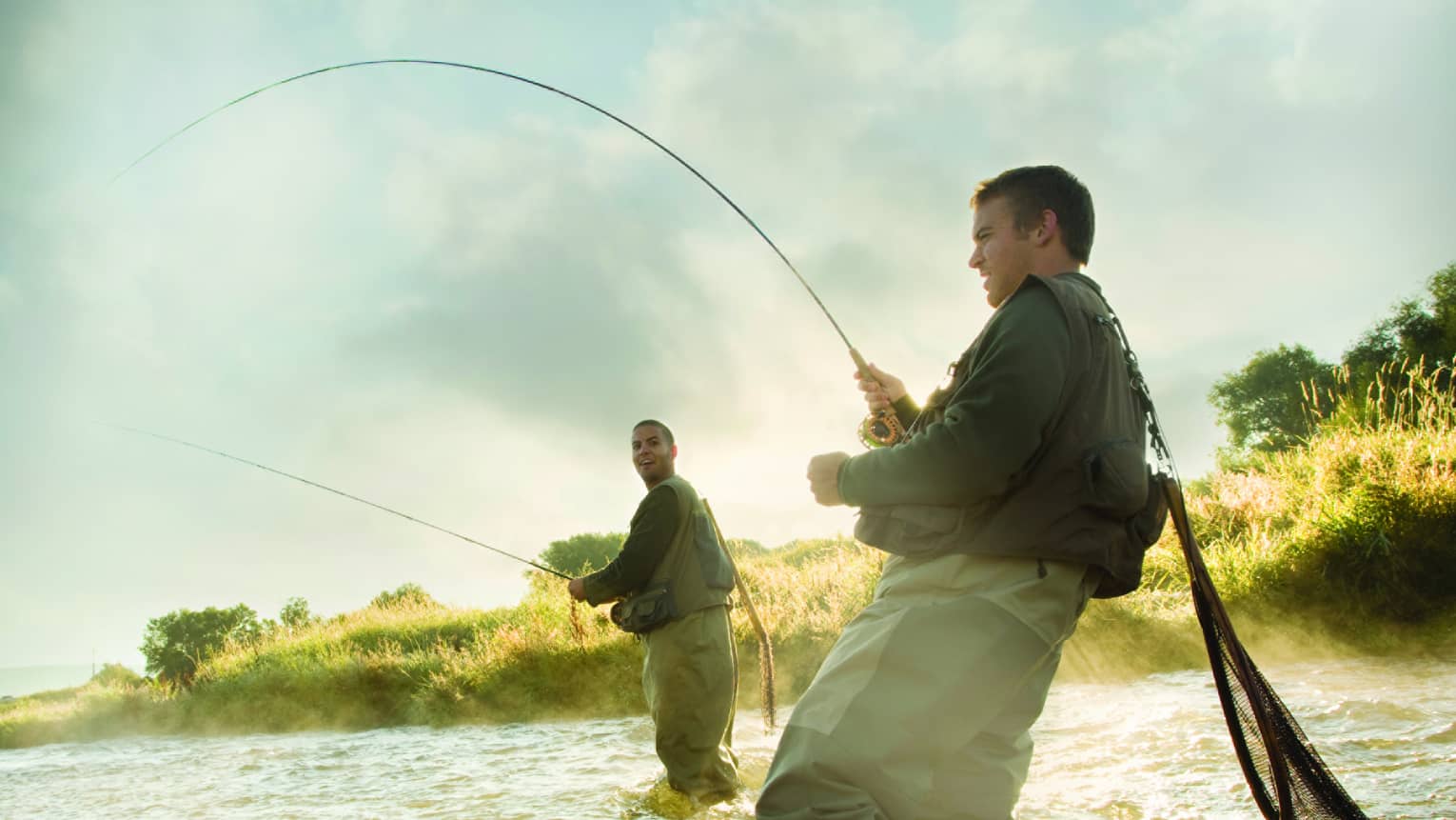 Two people fly fishing in a river, standing knee-deep in the water, surrounded by mist and greenery.