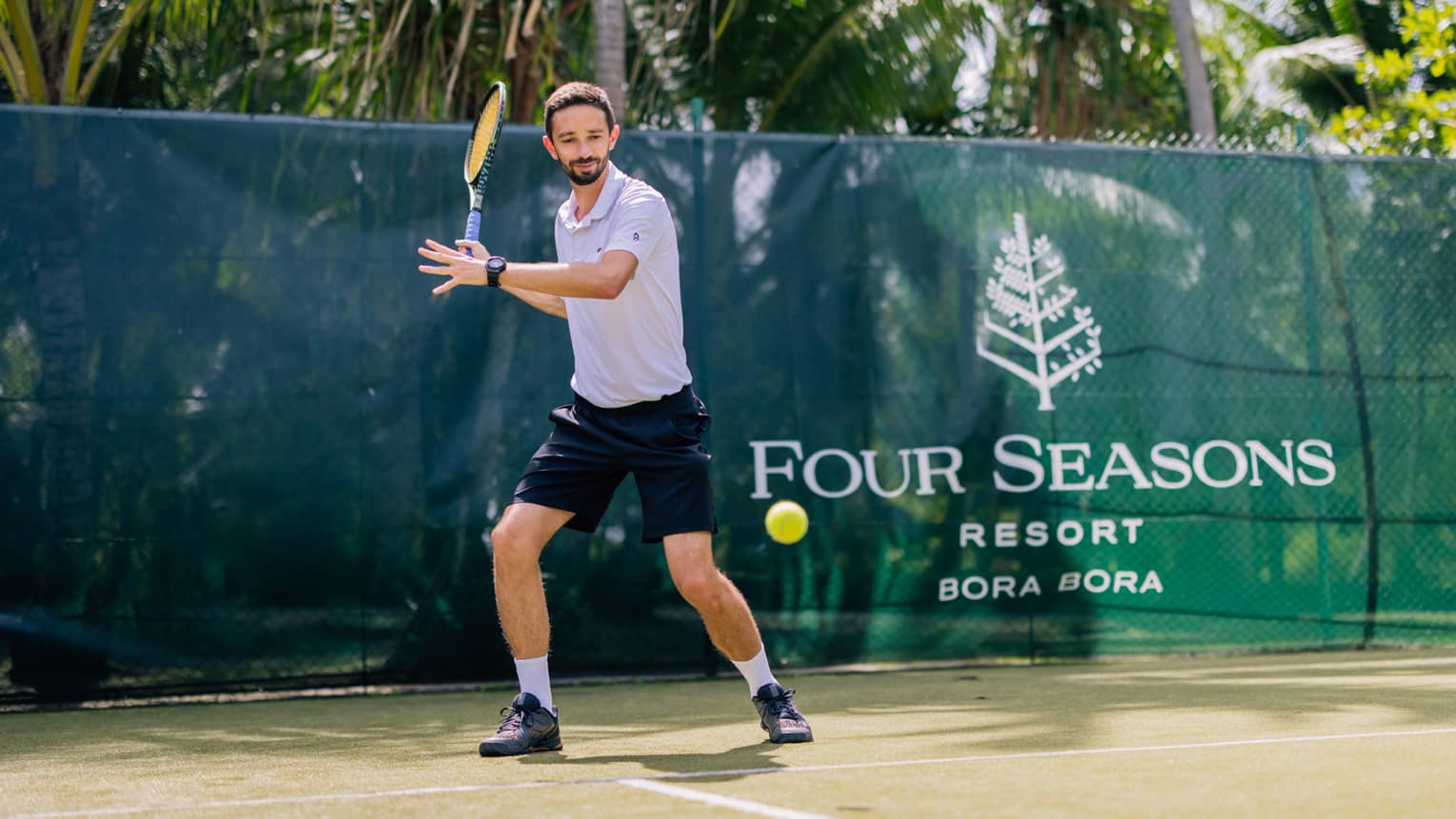Standing behind the baseline on a court surrounded by palm trees, a tennis player prepares to hit a forehand.