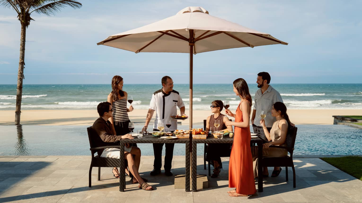 Friends gathered at a table with food setup and view of pool and beach in background