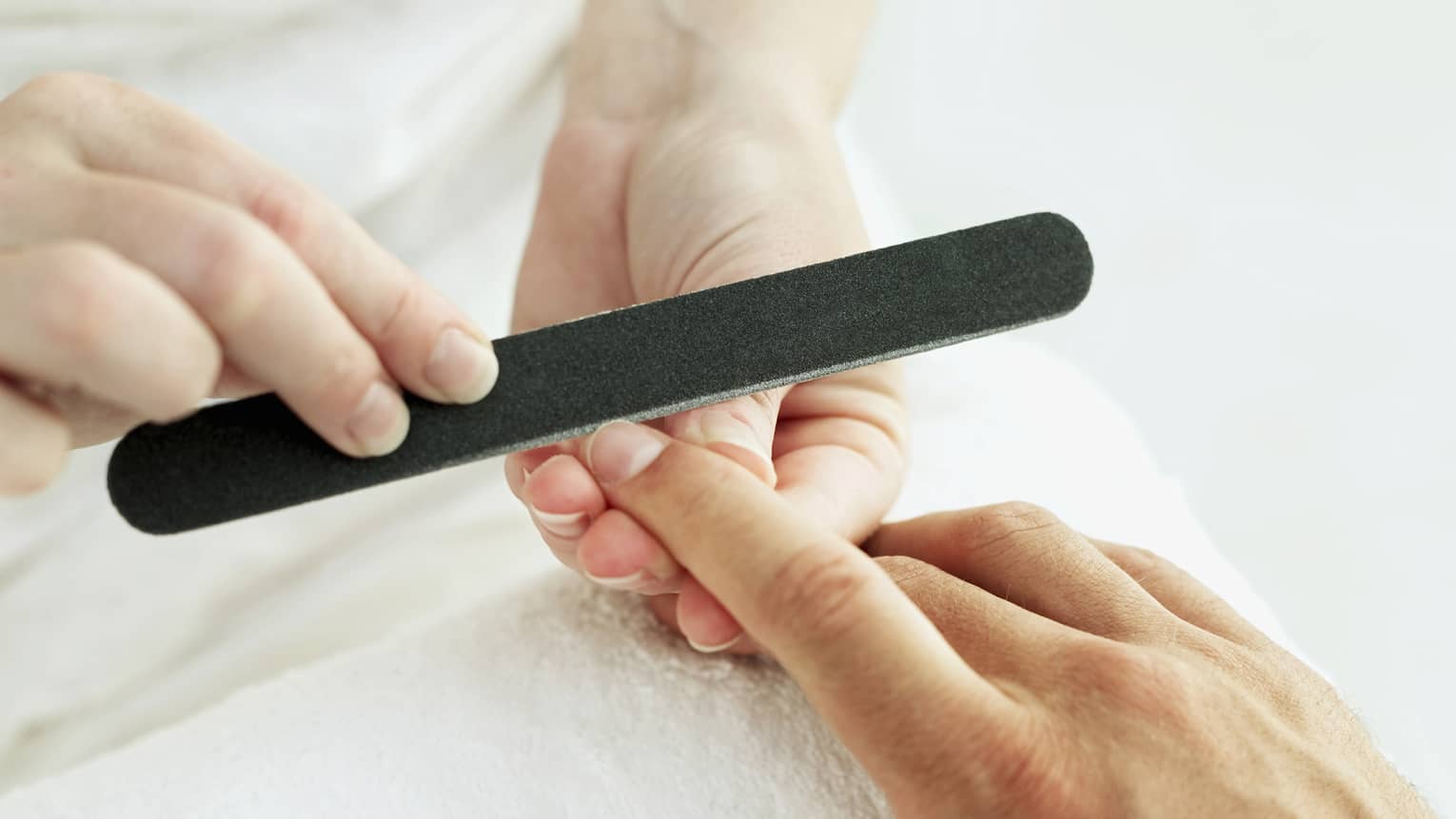 Woman holds manicure nail file to man's fingernails over white towel