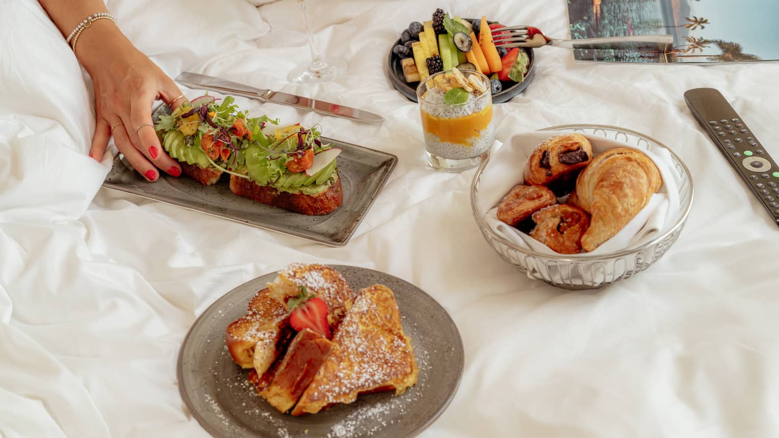 A few small plates of food items like pastries and French toast are arranged on a bed, next to a magazine and remote control. A person's hand reaches for one of the food items.