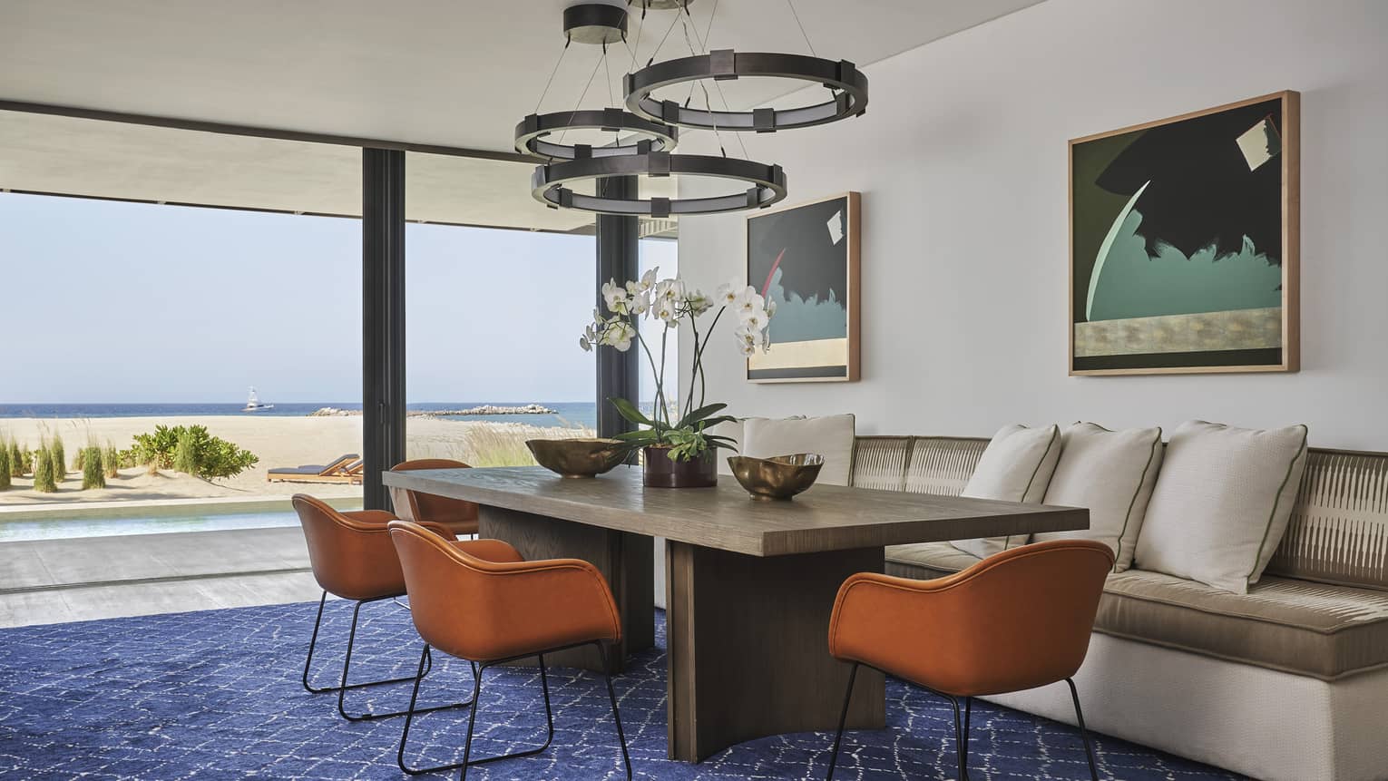Two Bedroom Sunrise Suite dining area with rectangular table, wrought iron overhead lighting, cream sofa, floor-to-ceiling windows with ocean views