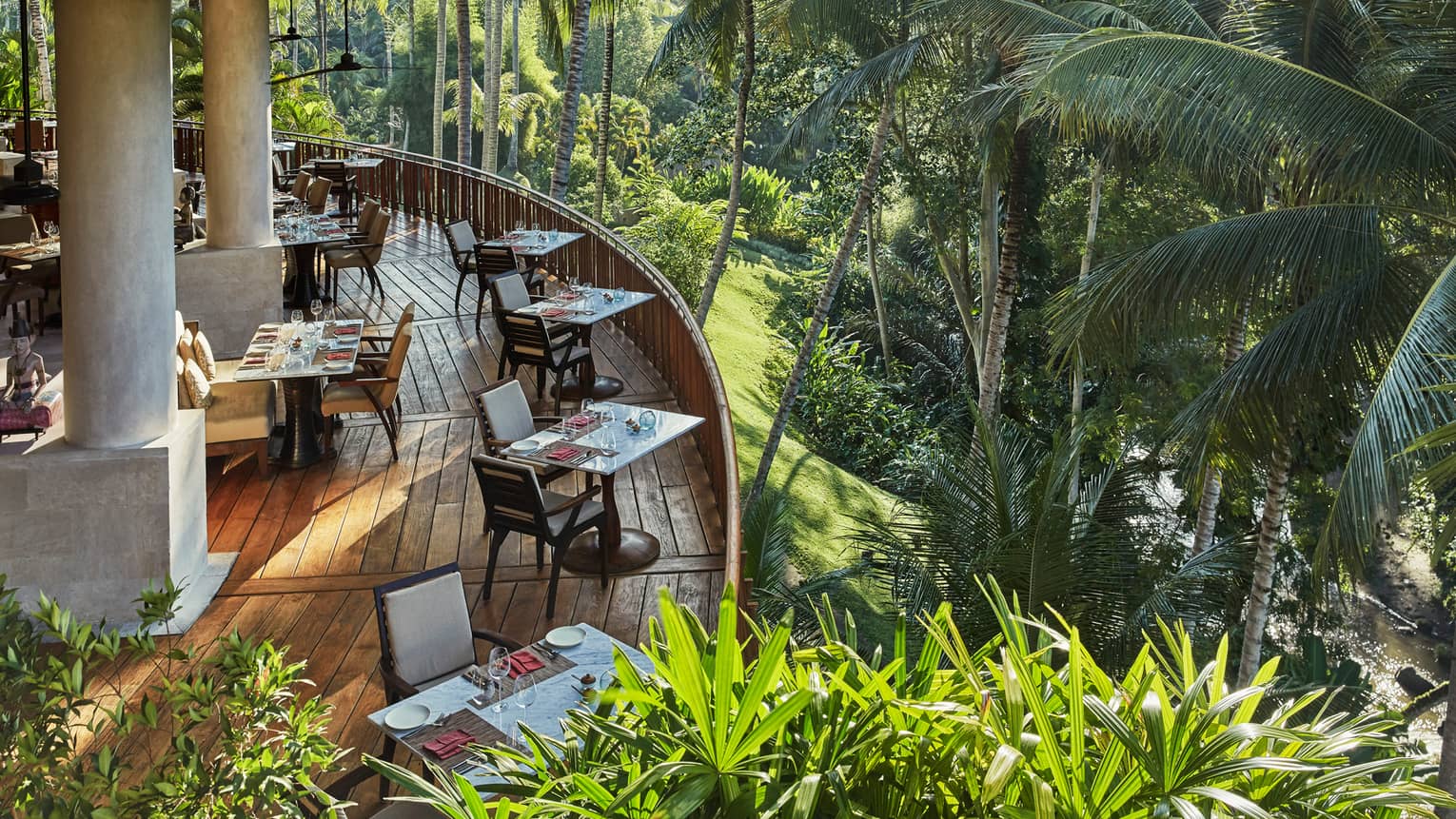 Looking down at curved wood patio of Ayung Terrace outdoor dining room, tables and chairs overlooking palm trees, forest