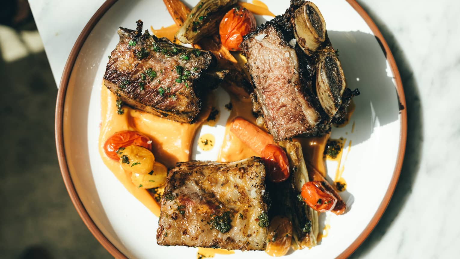 Three pieces of braised short rib served with roasted tomatoes and vegetables on a round white plate