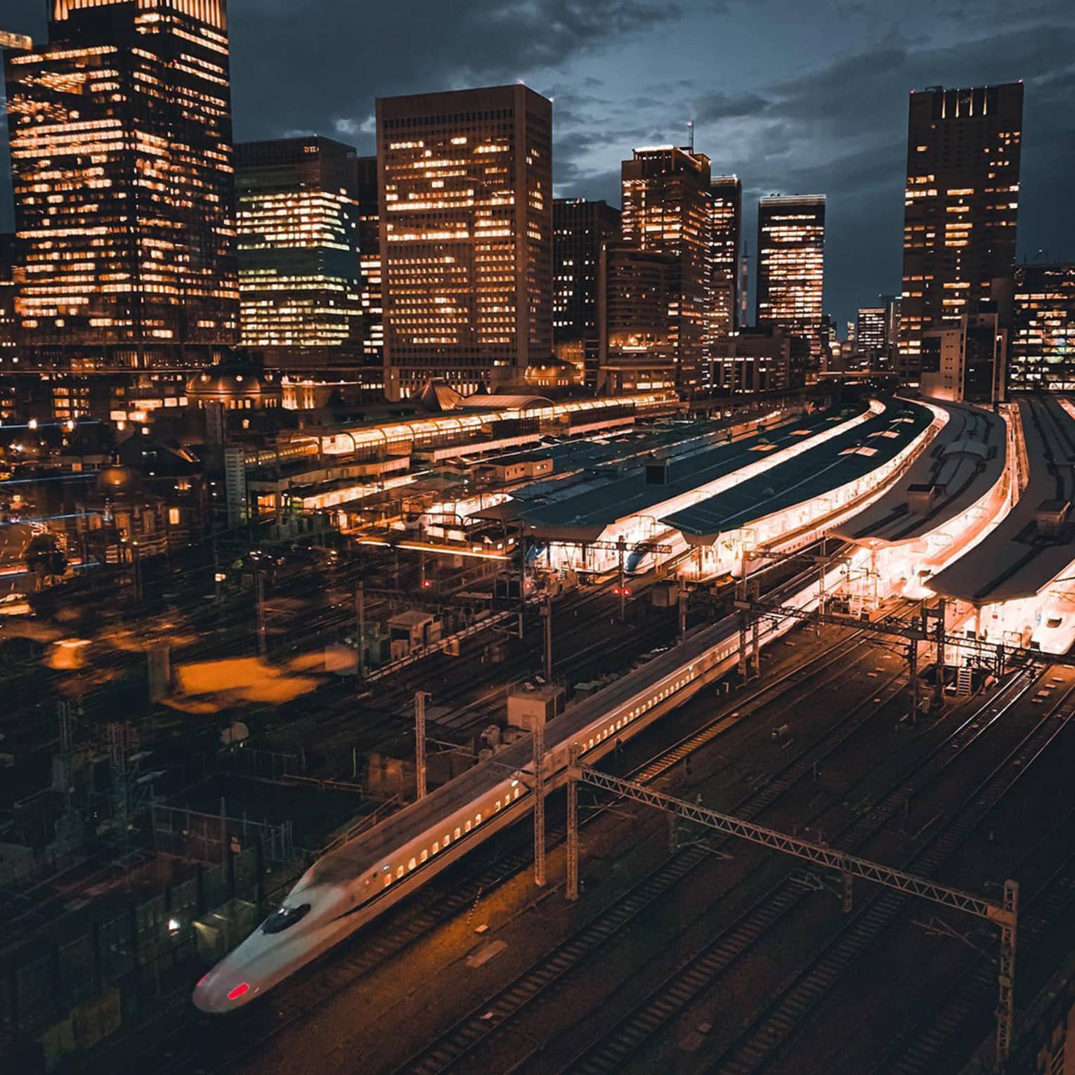 High-speed train passing through a city railway station at night with illuminated skyscrapers in the background