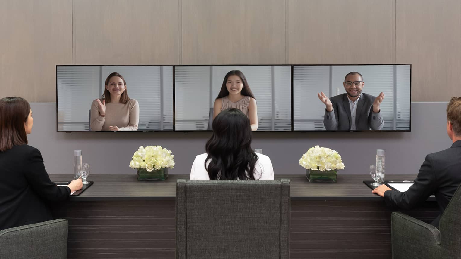 Several people in business clothes sit in front of a series of screens in a virtual meeting.