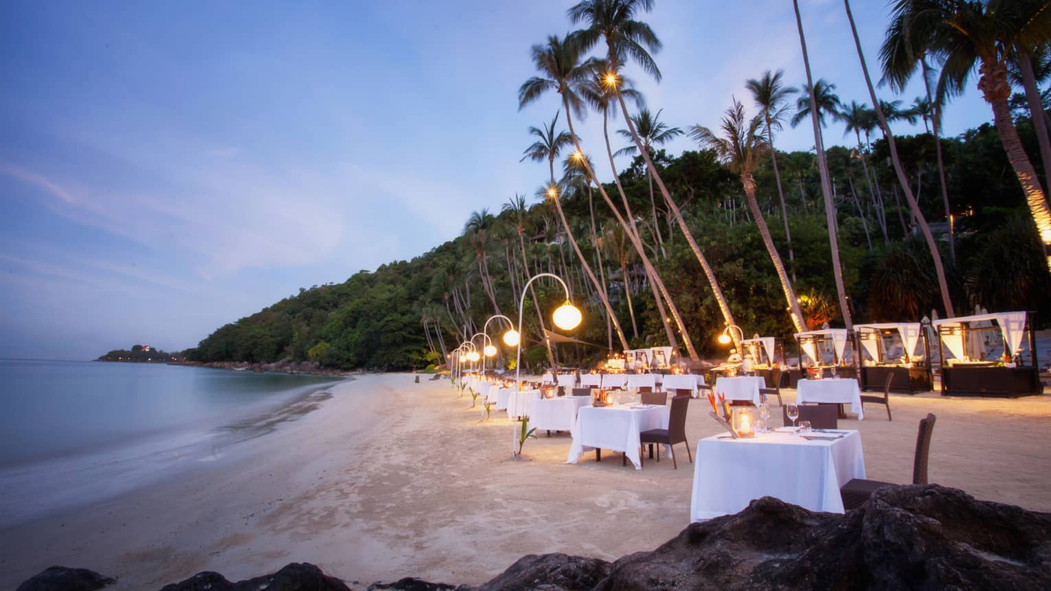 Elegant beachside dining setup at dusk with white-clothed tables, glowing lanterns and tall palm trees