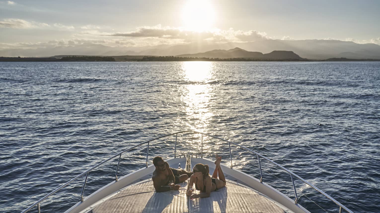 Couple wearing swimsuits relaxes on front yacht deck as sun sets over ocean