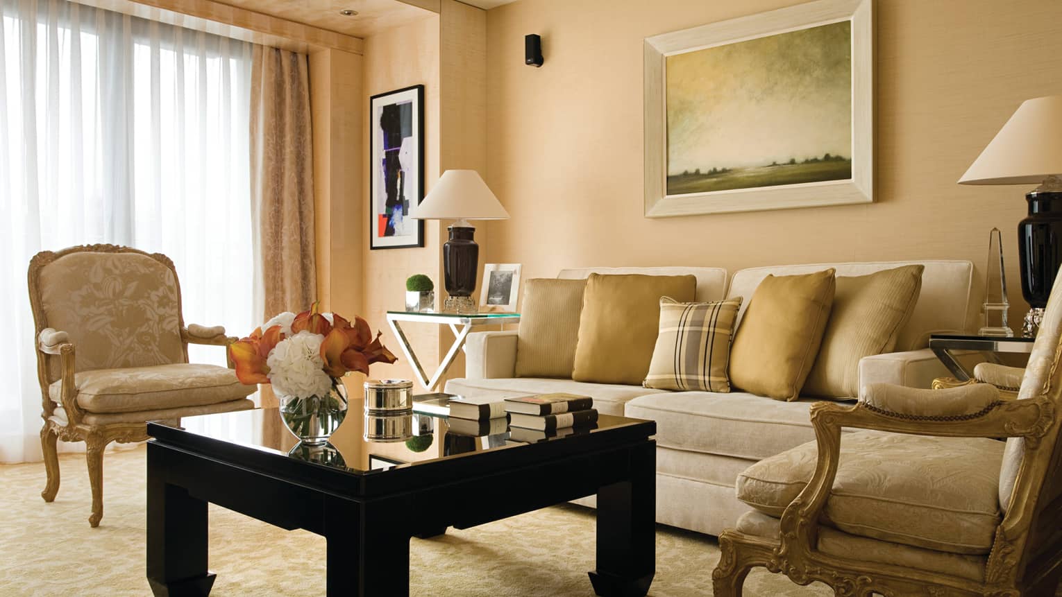 The Deluxe Suite at Four Seasons London Park Lane in Mayfair showcases a classic and luxurious living room setting. The room features a palette of soft beige and gold tones, with a plush beige sofa adorned with gold cushions. Two ornate, antique-style armchairs flank a sleek black coffee table, creating a sophisticated and inviting conversation area. Large windows are draped with heavy curtains, allowing natural light to enhance the warm atmosphere. The decor is completed by a traditional landscape painting above the sofa and elegant table lamps.