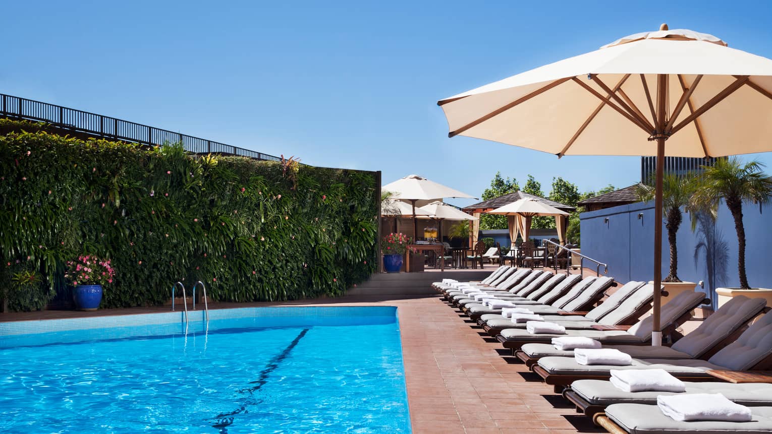White lounge chairs lined up on deck under patio umbrella by blue outdoor swimming pool