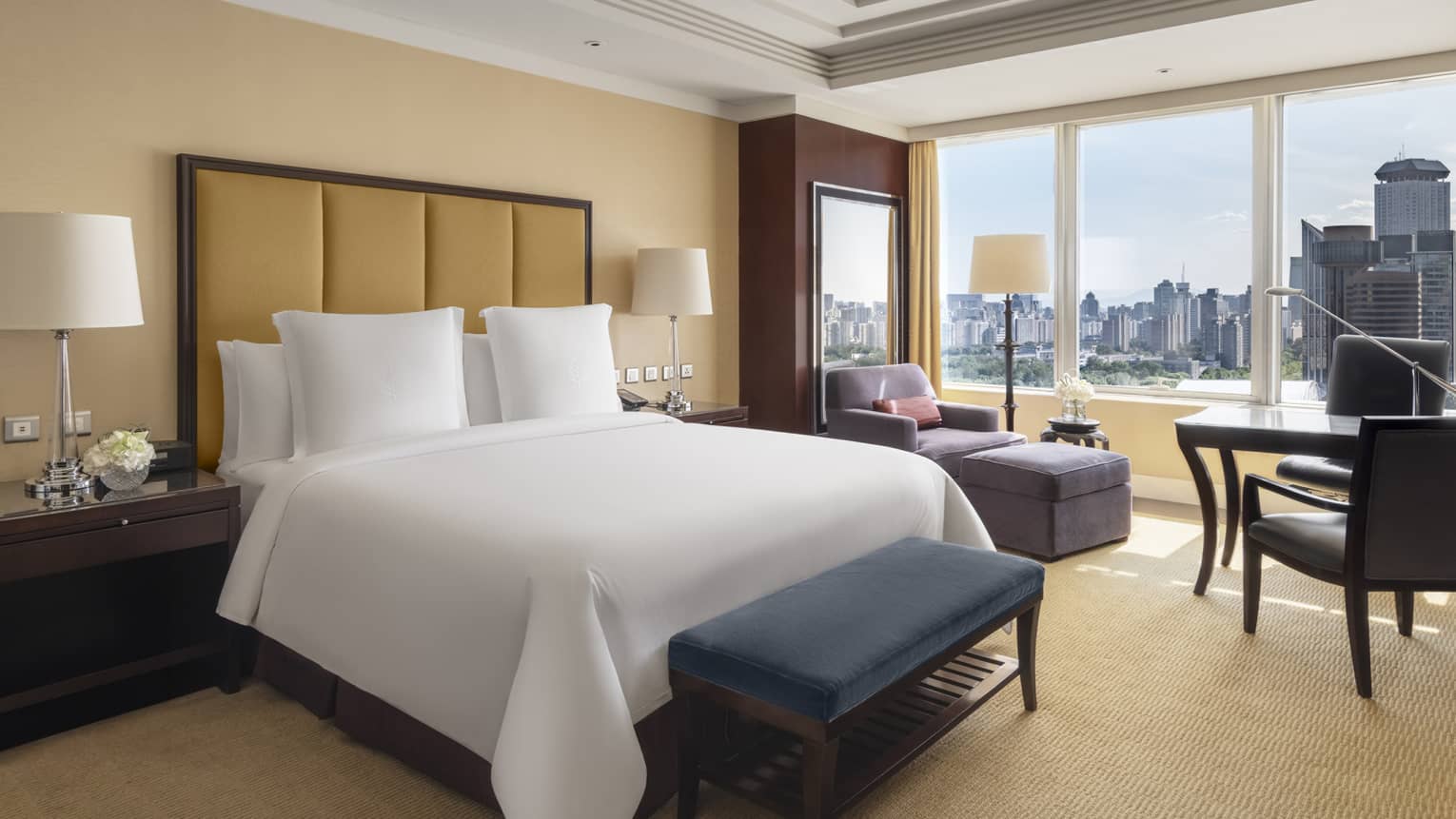 Hotel room with king bed, headboard, dark blue cushioned bench, windowside table and chairs