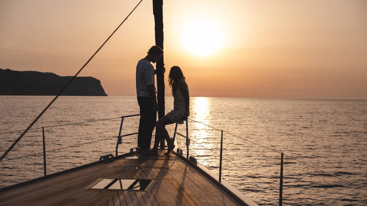 A couple at the pointed bow of a boat is silhouetted against the warm glow of the sun setting over calm, rippling water.