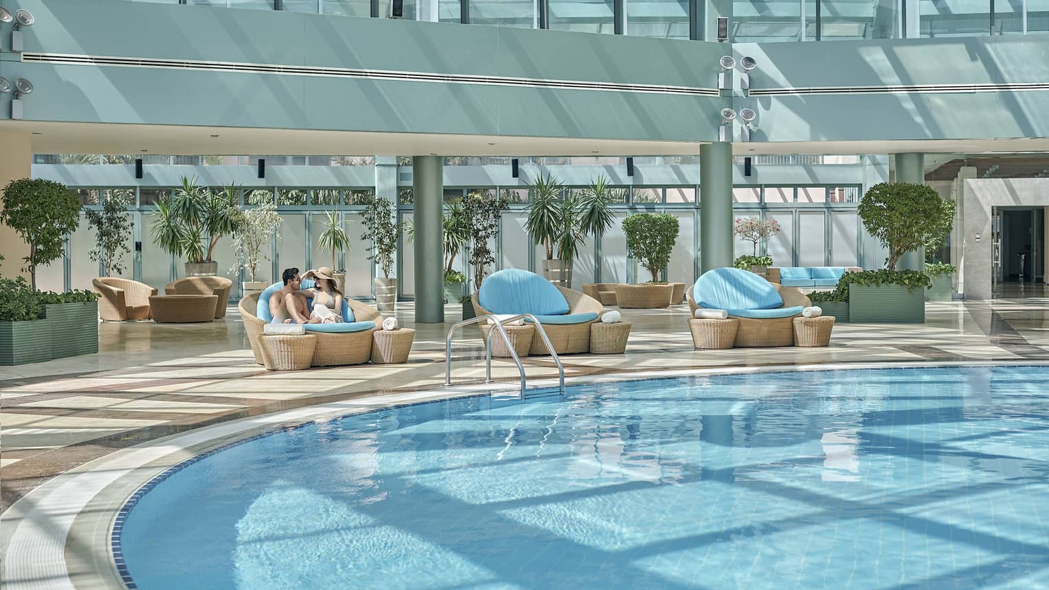 Couples sits on wicker patio chair on sunny deck by large outdoor swimming pool