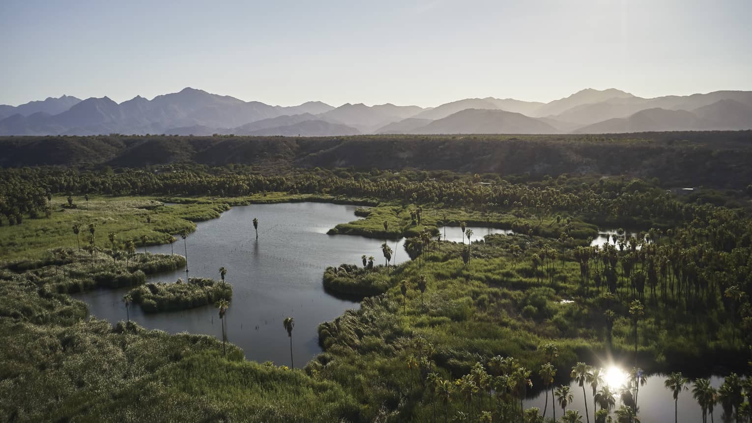 A marsh looking area with bonds and small patches of grass, mountains can be seen in the distance.