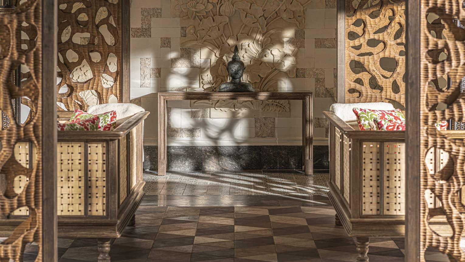 Dappled light dances over a stone bust and earth-tone tiles in an empty lounge with two couches facing opposite directions.
