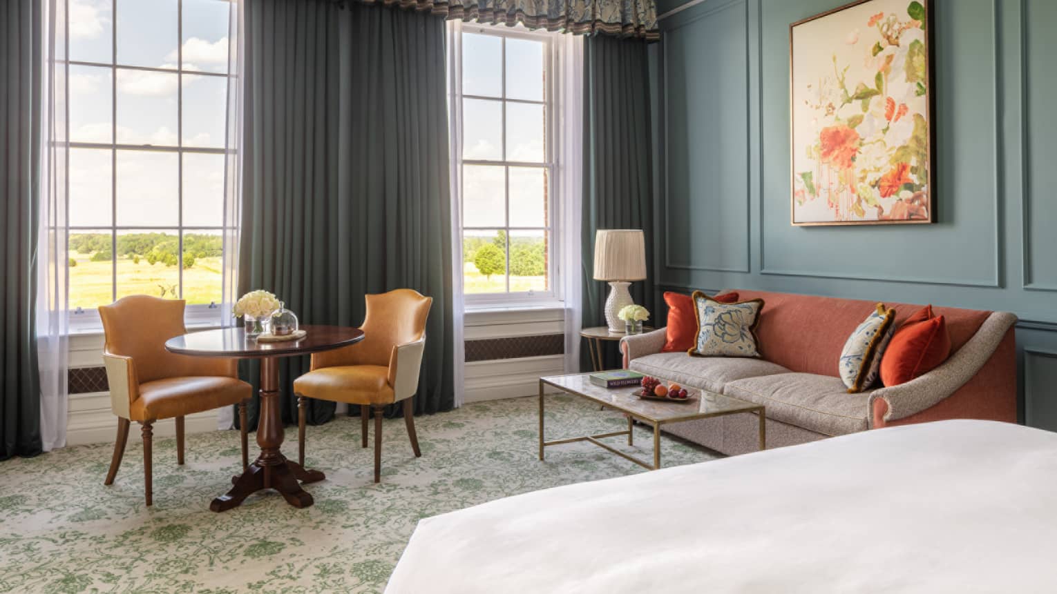 Guest room with bed, white linens, paisley carpeting, red and cream couch, small table for two, turquoise walls, twin windows with blue drapes